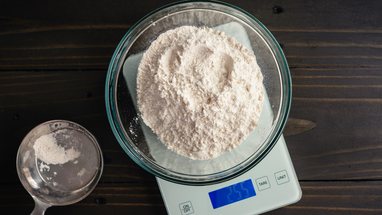 A glass bowl of flour on digital scale next to measuring cup