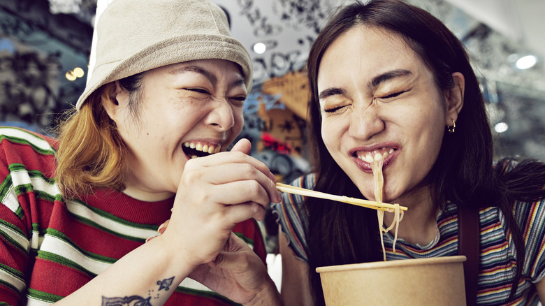 Happy young woman feeding noodles with chopsticks to female friend at street