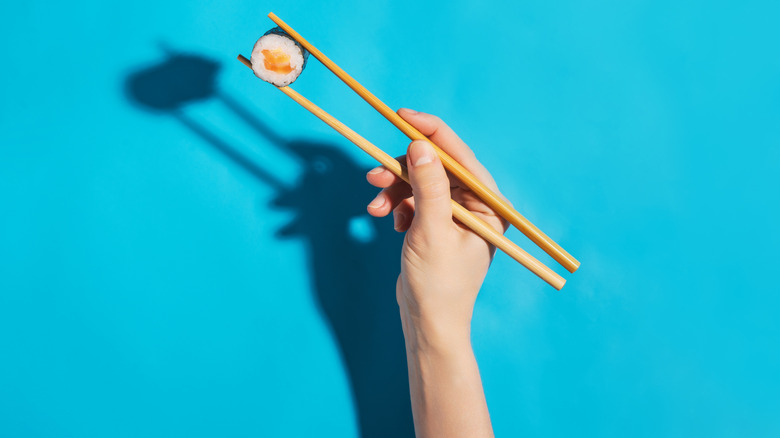 Hand with chopsticks holding salmon maki sushi roll on a blue background. Brightly lit studio shot.