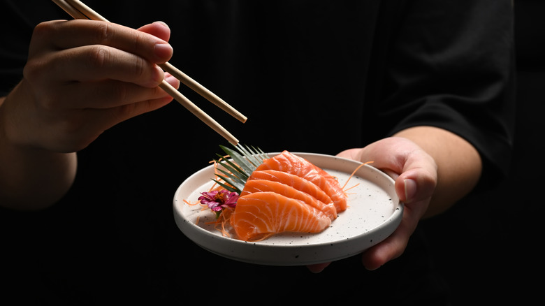 Man holding salmon sashimi with chopsticks on black background.