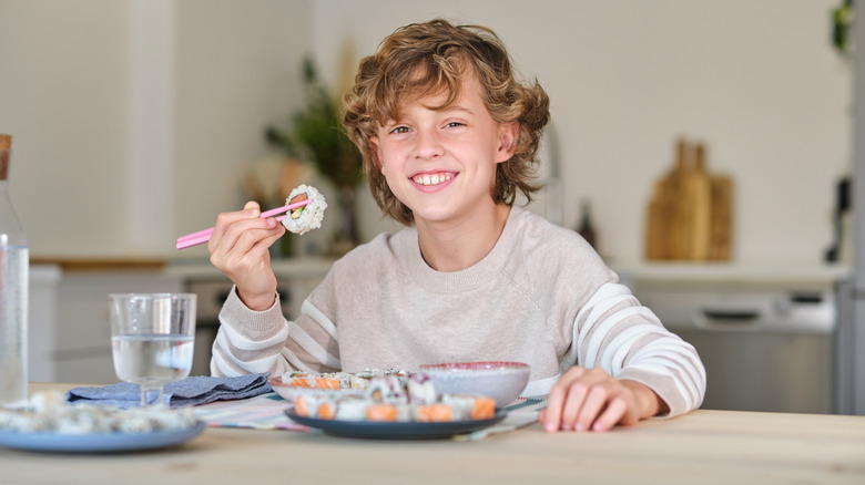 Smiling boy sitting at wooden table and looking at camera while eating tasty sushi rolls with chopsticks in dining room at home