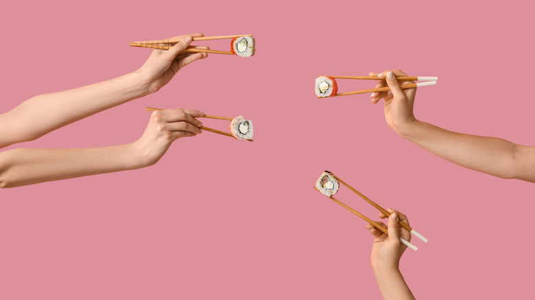 Four hands holding chopsticks with different tasty sushi rolls on pink background
