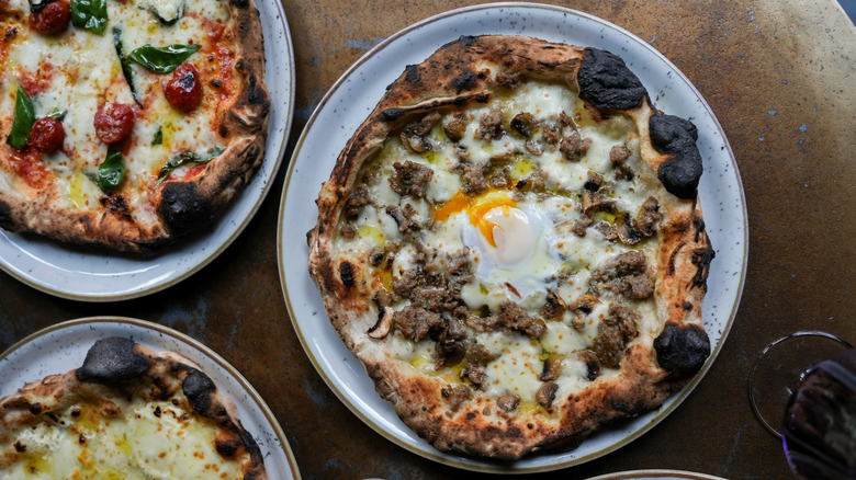 A series of pizzas on speckled white plates, all displaying charred air bubbles and a pizza with a runny egg in the middle, surrounded by some beef and mushrooms