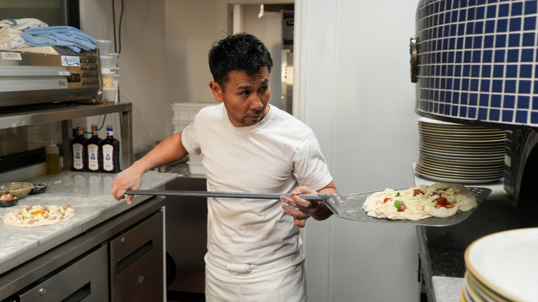 Tamaki, wearing a white T-shirt and apron, slides a pizza on a metal peel into a blue-tiled oven