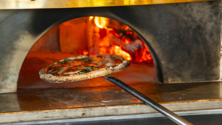 A Neapolitan pizza on a metal peel being inserted into a wood-burning oven