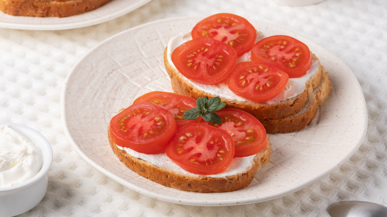 two open-faced tomato sandwiches on a white plate, one garnished with a small sprig of oregano