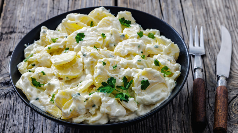 A bowl of creamy potato salad sits on a wooden table with a knife and fork laid beside it