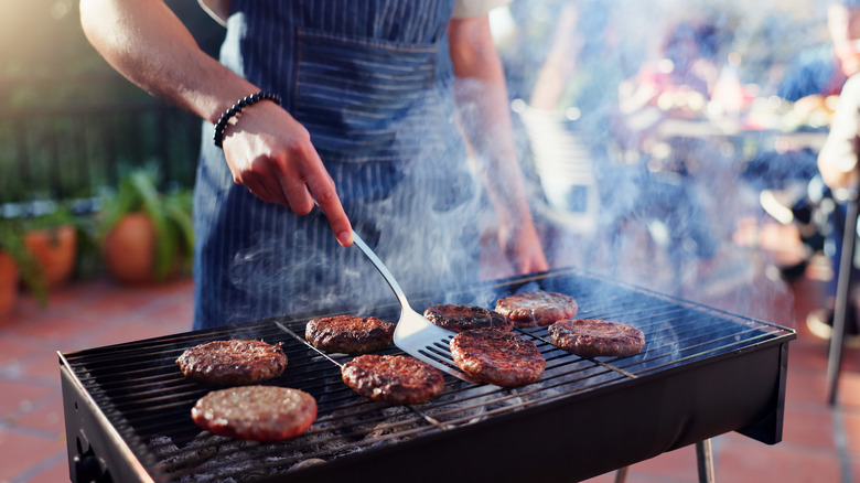 A man in an apron stands over an outdoor grill cooking eight burgers, preparing to flip one with a metal spatula