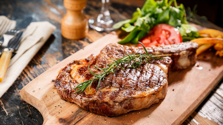 A cooked ribeye tomahawak steak rests on a cutting board on a wooden table.