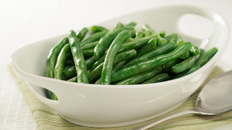 a white ceramic dish of cooked green beans with a serving spoon beside it resting on a tea towel