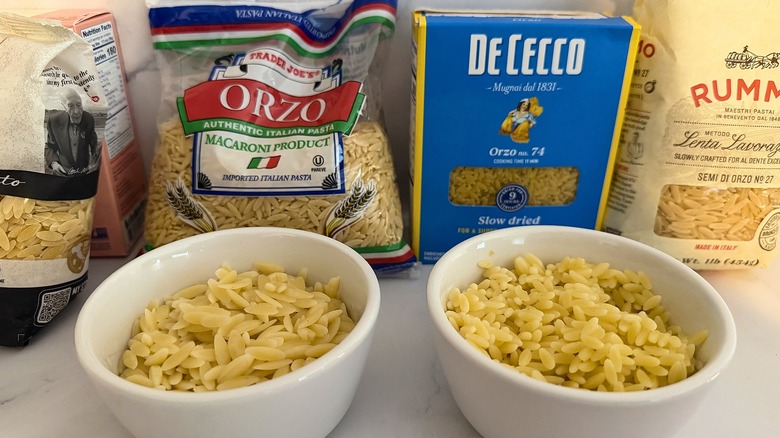 Two white cups of cooked orzo side by side, orzo boxes and bags in background