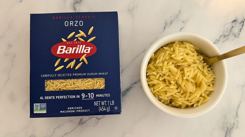 Package of Barilla orzo next to small cup of orzo with spoon, all on marble surface