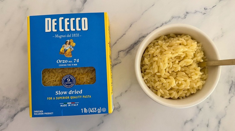 Package of De Cecco orzo next to small cup of orzo with spoon, all on marble surface