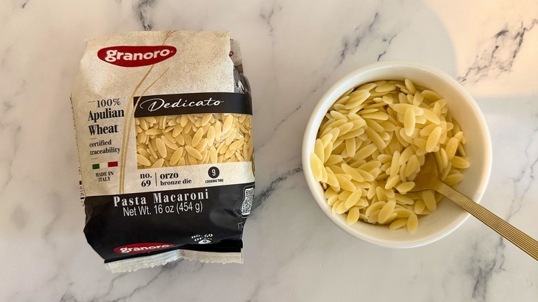 Package of Granoro orzo next to small cup of orzo with spoon, all on marble surface