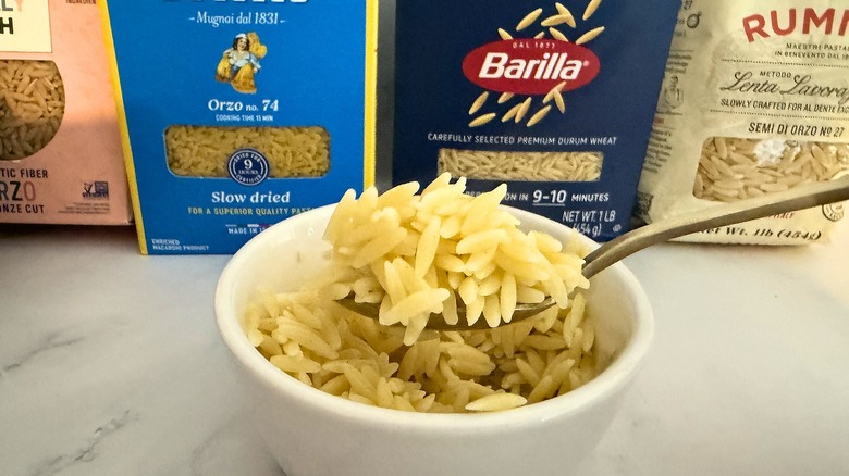 Spoon holding orzo over orzo over small cup of orzo, packages of orzo in background