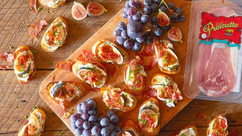 Serving board with sliced baguette piled with cheese and prosciutto, grapes and package of prosciutto beside
