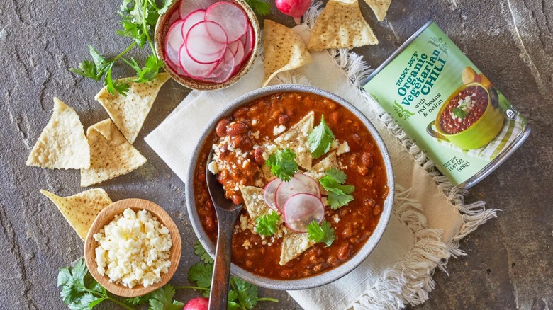 A bowl of Trader Joe's Vegetarian Chili next to a can of the chili, radishes, chips, cheese, and herbs