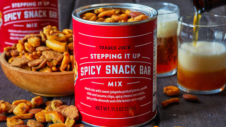 A tin of Trader Joe's Stepping it Up Spicy Snack Bar Mix open on a table next to a bowl of the mix and two glasses of beer