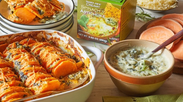 Trader Joe's creamy spinach and artichoke dip box on a table with the actual dip in a ceramic bowl, a casserole dish with sweet potatoes, a plate of sweet potatoes, a bowl of sweet potatoes, and some parmesan cheese.