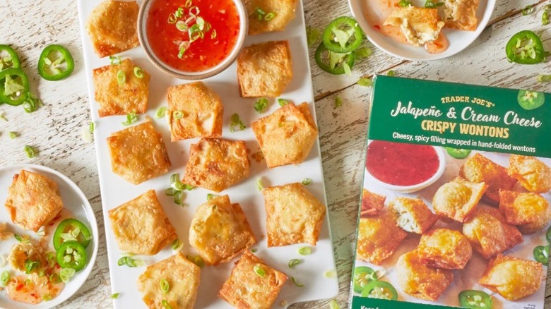 A box of Trader Joe's jalapeno and cream cheese crispy wontons sitting beside a platter of finished wontons and red sauce, as jalapeno slices are scattered about.
