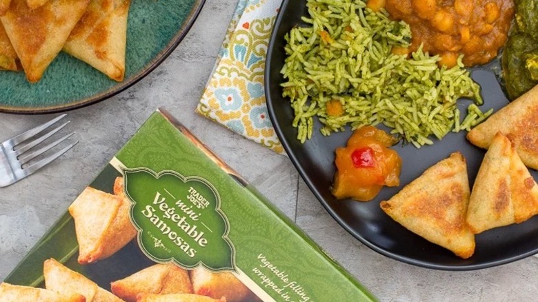 A box of Trader joe's mini vegetable samosas on a marble table with two plates, one teal and one black, with baked samosa's, sauce, and rice.