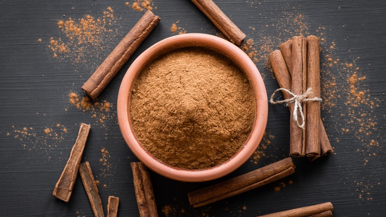 Small bowl of ground cinnamon surrounded by whole cinnamon sticks on dark surface