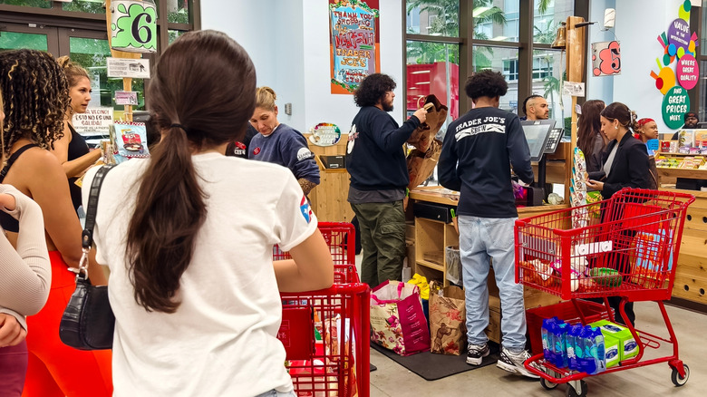 Checkout area in Trader Joe's store with cashiers behind wooden counters while customers stand in line with red shopping carts