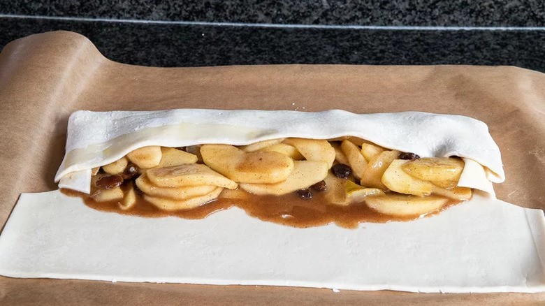 Uncooked and partially folded apple strudel on a sheet of parchment paper