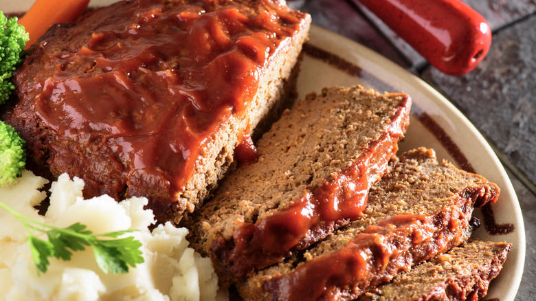 A sliced, ketchup-glazed meatloaf on a plate with some vegetables, herbs, and mashed potatoes.