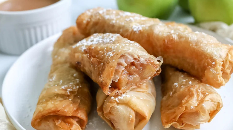 A stack of freshly made apple pie phyllo rolls on a plate, dusted with powdered sugar, with several apples and a bowl of caramel dipping sauce in the background