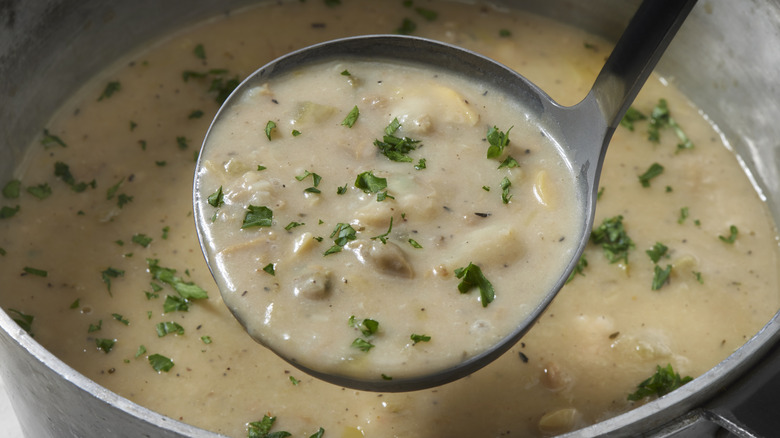 a ladle full of clam chowder topped with fresh herbs above a pot of the same chowder.