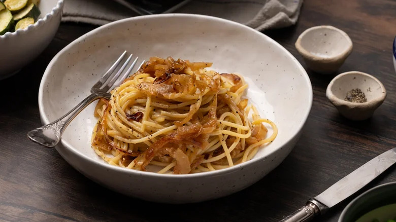A bowl of simple caramelized onion pasta with a fork on a table with little dishes of salt and pepper