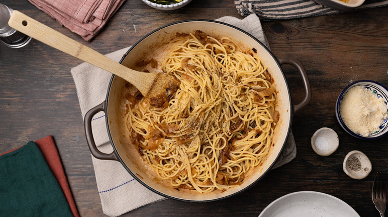 A pot of caramelized onion pasta with a wooden spoon on top of a table with parmesan, salt, and pepper