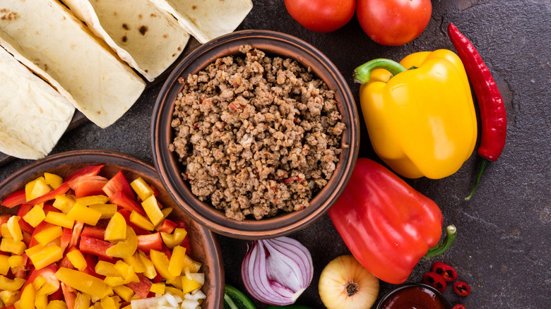 A flat surface covered with taco ingredients, including tortillas, a bowl of chopped veggies, a bowl of cooked ground beef, and various whole vegetables, including bell peppers, chilies, and onions.