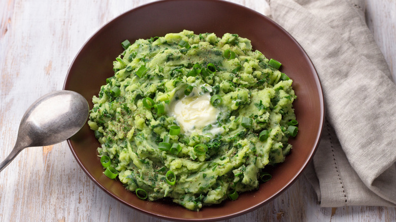 A bowl of creamy green colcannon with a melting pat on butter in the center and a spoon resting on the edge