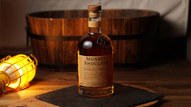 A bottle of Monkey Shoulder blended Scotch whisky sits on a mat, on a wooden surface, with an illuminated electric lamp lying beside it and a wooden tub in the background