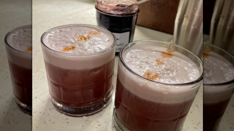 Two freshly prepared Monkey Jam Sour cocktails in rock glasses, sitting on a white surface with an open pot of jam in the background