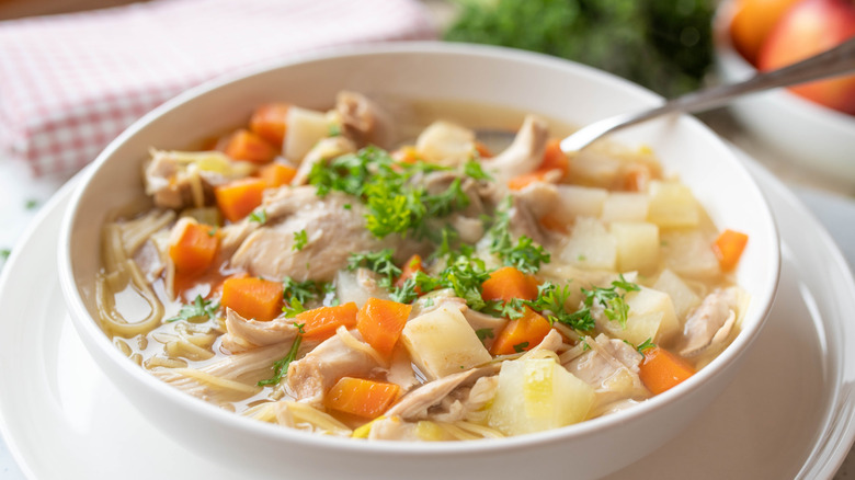 bowl of chicken noodle soup with parsley and carrots and meatballs visible, sitting on a folded towel next to some parsley on a table top