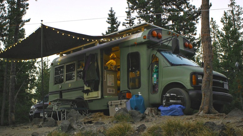a converted short bus parked in the forest with a lit awning outside and someone standing inside the open door