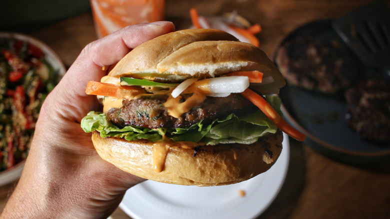 a hand holding a banh mi burger topped with lettuce, jalapeños, and pickled carrots and daikon