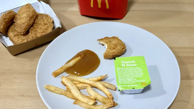 A white plate featuring a container of McDonald's sweet and sour sauce alongside some french fries and a partially eaten Chicken McNugget. There are more McNuggets and fries in the background