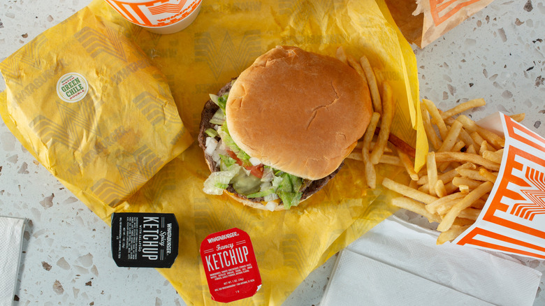Burger from Whataburger on yellow paper wrapping next to ketchup dip packets and cardboard sleeves of French fries