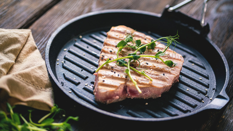 a grilled tuna steak topped with sesame seeds and microgreens in a cast iron grill pan on a wooden table