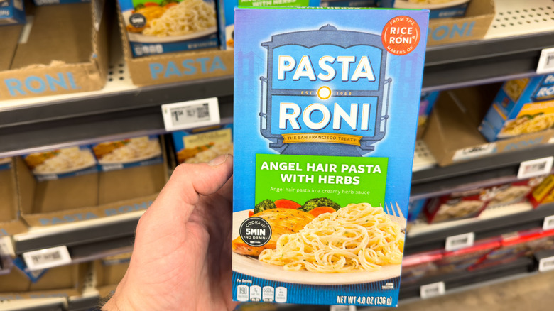 A person holding a box of Pasta Roni angel hair pasta with herbs flavor in front of a grocery store shelf