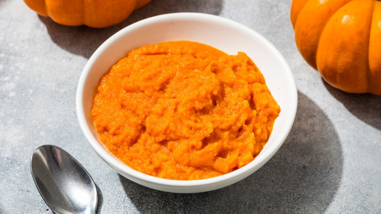 A white bowl full of pumpkin puree on a table next to two small pumpkins