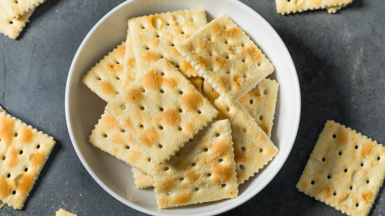 A bowl of saltine crackers against scattered saltines on gray table