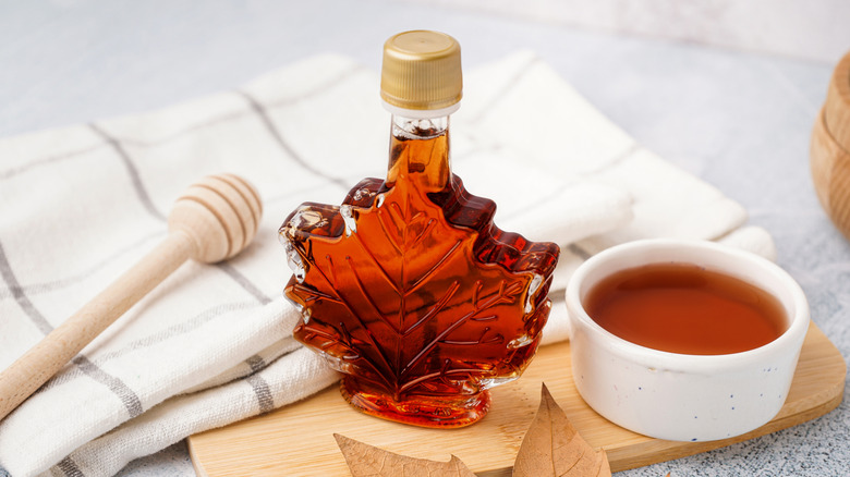 A bowl of maple syrup next to a leaf-shaped bottle, honey dipper, and tea towel