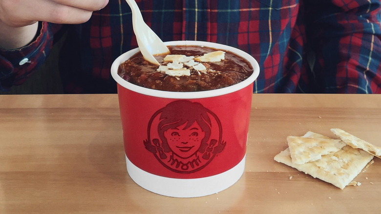 A person in a plaid shirt dipping a spoon into a red cup of Wendy's chili with crackers crushed on top next to crackers on table