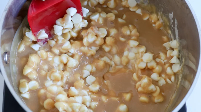 A pot of white chocolate chips in light brown liquid being stirred with a red silicone spatula