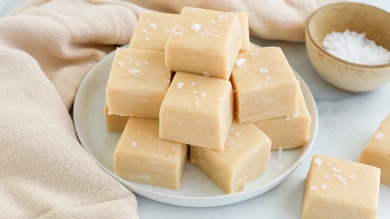 Salted caramel fudge stacked on a small plate beside a napkin and small wooden bowl of coarse salt.